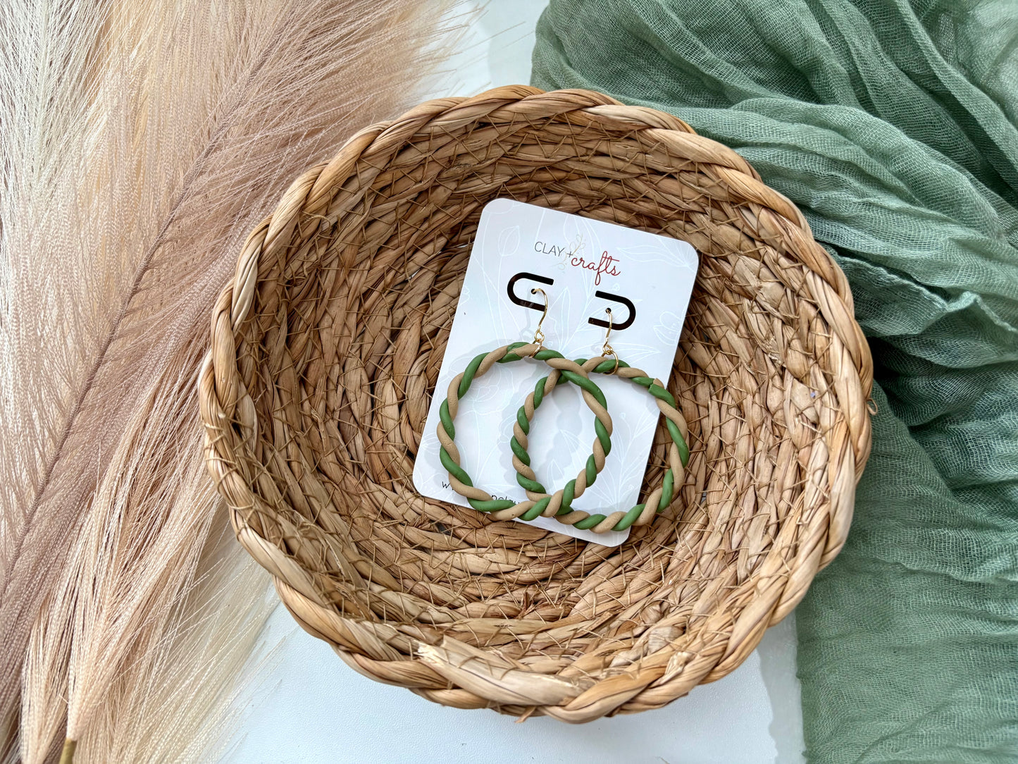 Pair of green and beige hoop earrings on a card with a wicker basket and green fabric in the background.