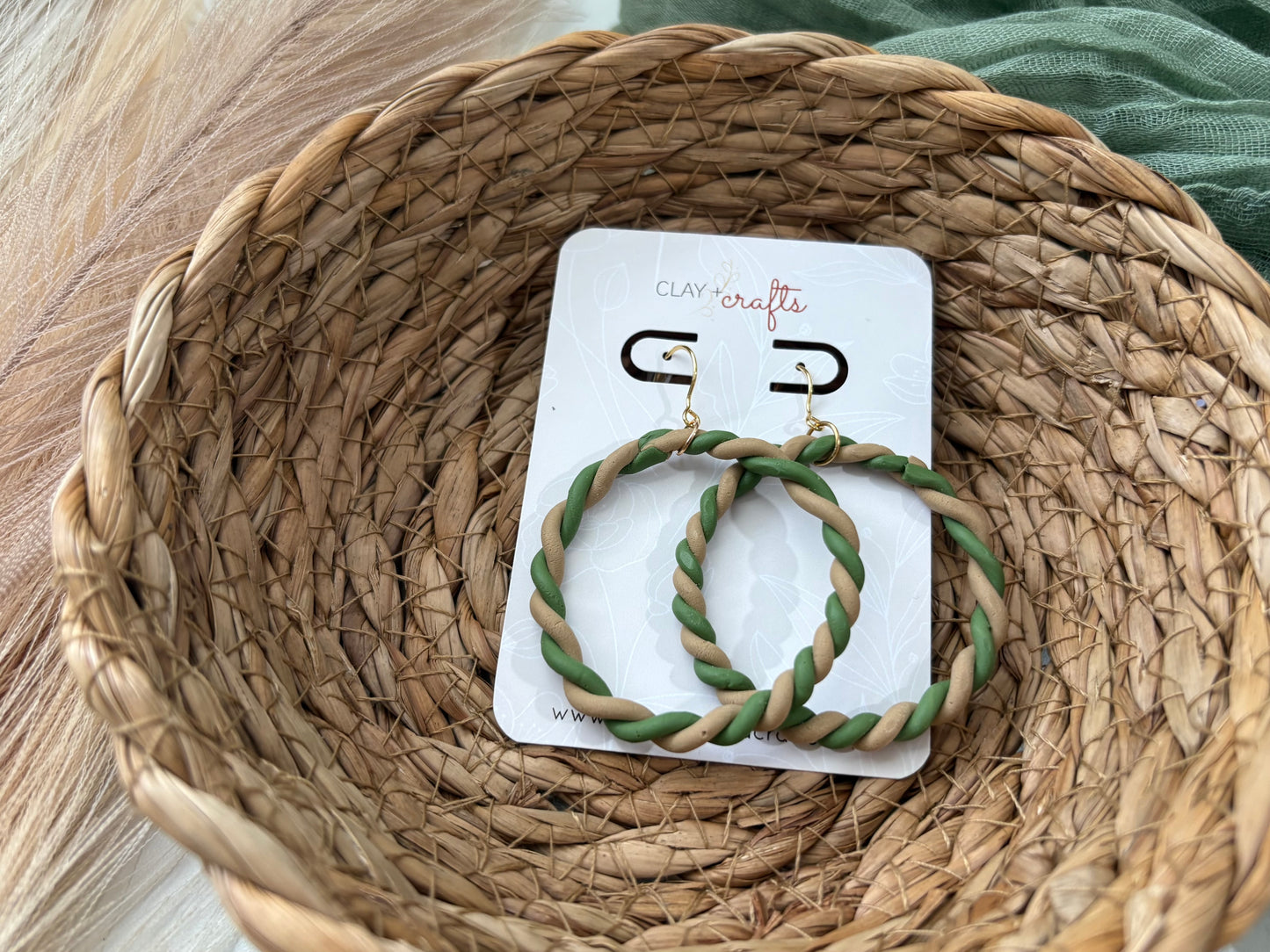 Green and beige circle braided hoop earrings on a Clay + Crafts brand card, placed on a woven surface
